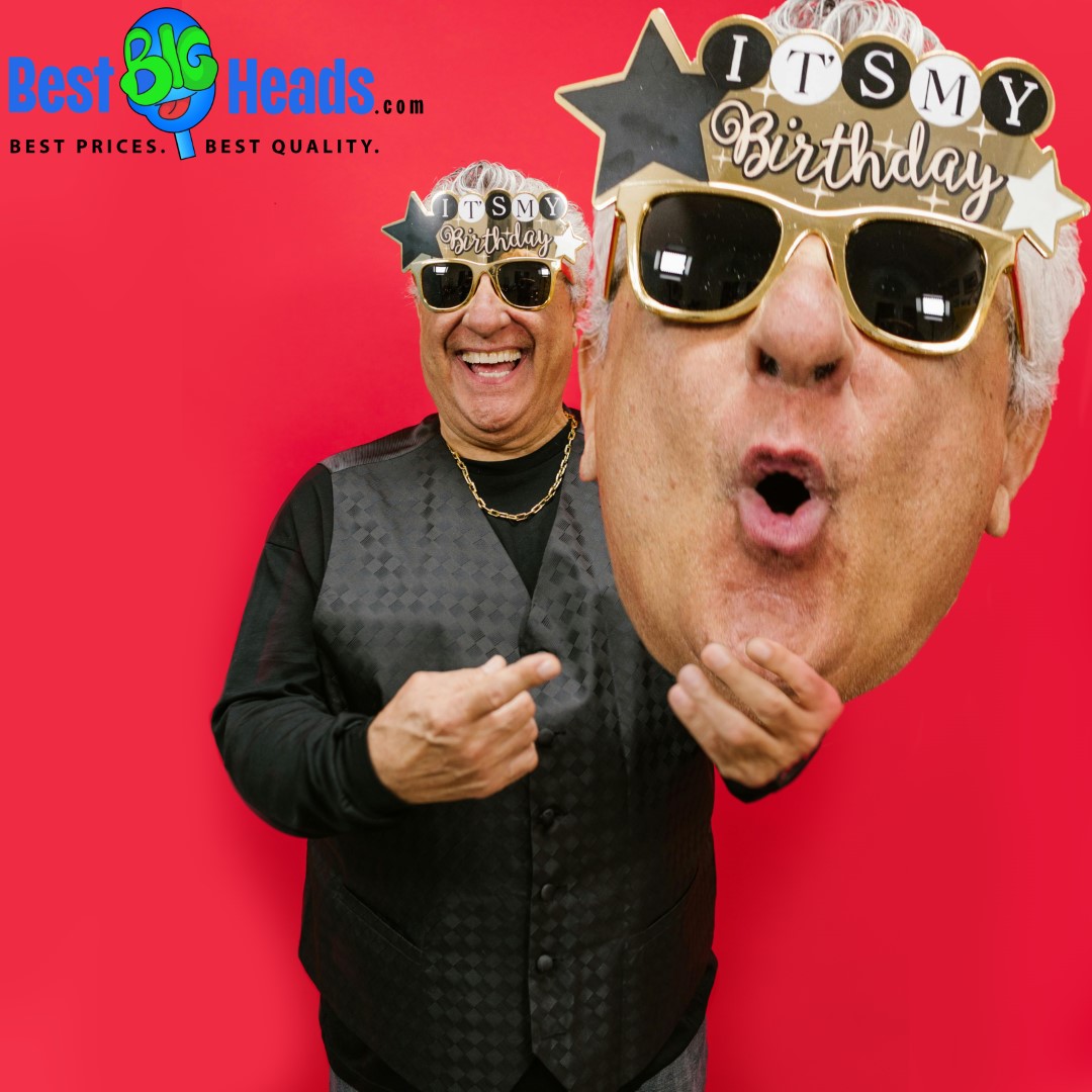 A man wearing festive "It's My Birthday" sunglasses, smiling and holding a giant head cutout of himself in front of a red background.